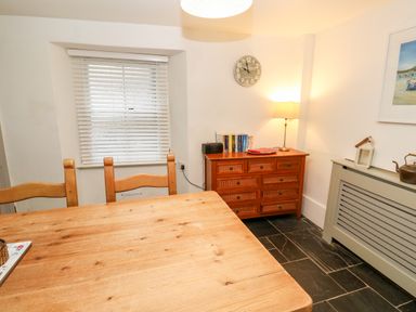 A dining room with wooden table and chairs a wooden dresser with books lamp and clock on the wall at Bay Tree Cottage in West Looe