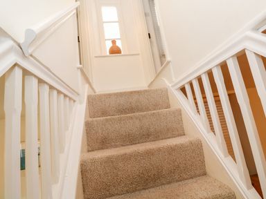 Carpeted staircase with white wooden railing and a window with a woven vase on the sill at Bay Tree Cottage in West Looe