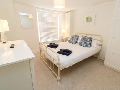 A bedroom with a bed with pillows and folded towels a dresser with a lamp and remote window with blinds at Bay Tree Cottage in West Looe