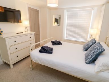 A bedroom with a white bed and blue pillows a white dresser with a lamp and a wall-mounted TV at Bay Tree Cottage in West Looe
