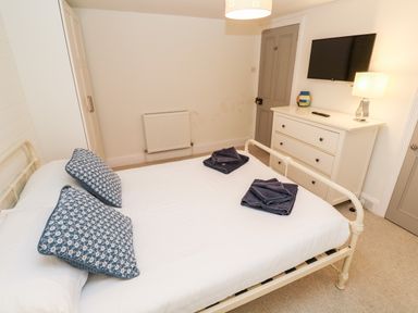 A bedroom with a white metal bed frame, blue patterned pillows, folded dark towels on the bed, a white dresser with a lamp and TV mounted above at Bay Tree Cottage in West Looe