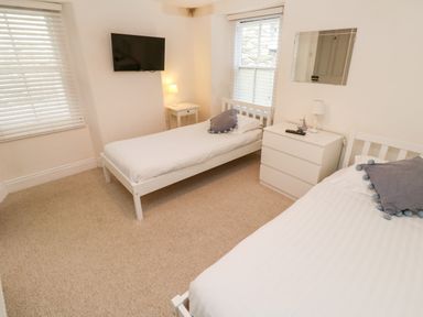 A bedroom with two single beds white furniture a wall mounted TV and windows with blinds at Bay Tree Cottage in West Looe