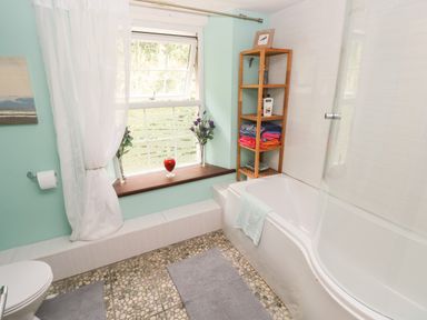 A bathroom with a bathtub a wooden shelf with towels and toiletries a window with white curtains and flowers at The Old Mill in Neyland near Milford Haven