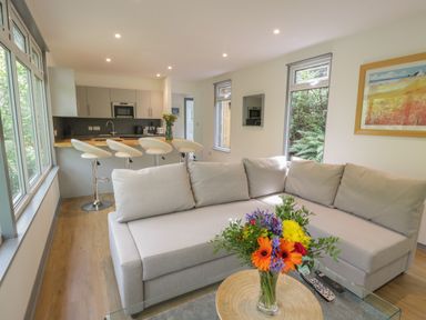 A living room with a gray sofa, a glass coffee table with flowers, a kitchen with stools, and windows at The River Lodge in Oban