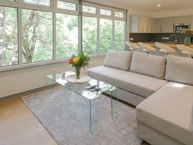 A living room with a beige sofa glass coffee table with flowers and a kitchen with bar stools in the background at The River Lodge in Oban