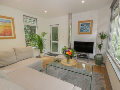 A living room with a beige couch glass coffee table flowers on the table tv and plants by the windows at The River Lodge in Oban