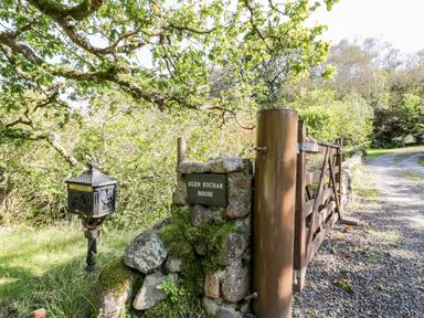 A wooden gate and stone pillar with a sign reading Glen Euchar House next to a mailbox by a gravel driveway surrounded by trees