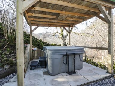 An outdoor hot tub with a cover and steps under a wooden shelter surrounded by trees at The River Lodge in Oban