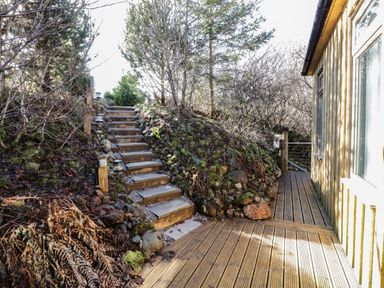 Outdoor wooden steps leading up a small hill with plants and trees next to a wooden deck beside a yellow building at The River Lodge in Oban