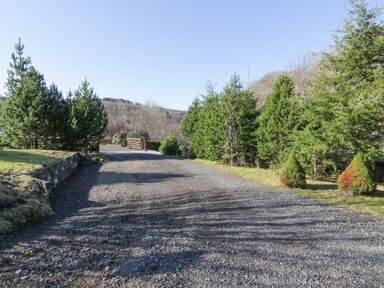 A gravel driveway bordered by evergreen trees leading to a wooden gate at The River Lodge in Oban