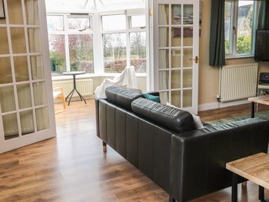 A living room with a black leather sofa wooden floors glass double doors and a small table with chairs near windows at The Shieling in Warkworth