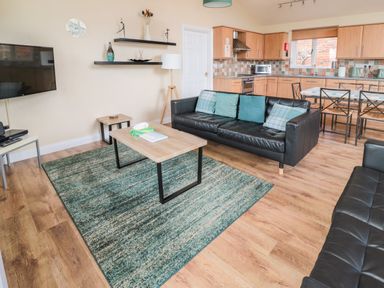 An open living room and kitchen with black leather sofas a wooden coffee table a patterned rug wood flooring and a dining table at The Shieling in Warkworth