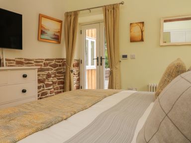 A bedroom with a bed a chest of drawers a wall-mounted TV curtains and a door leading outside at Langdale Barn in Sampford Courtenay
