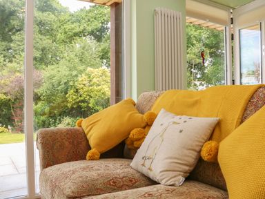 A patterned sofa with yellow cushions and a cushion with a duck illustration near large windows showing green trees outside at Langdale Barn in Sampford Courtenay