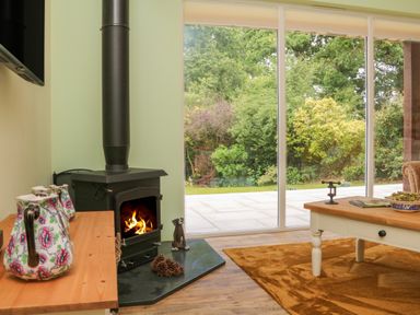 A living room with a wood burning stove next to a window overlooking a garden at Langdale Barn in Sampford Courtenay