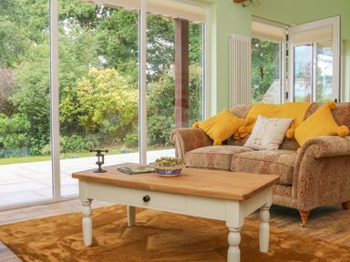 A living room with a patterned sofa with yellow pillows wooden coffee table and large glass doors showing garden at Langdale Barn Sampford Courtenay