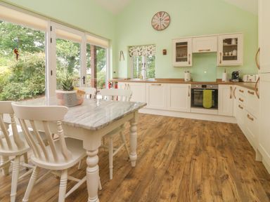 A kitchen with white cabinets a wooden floor a dining table with chairs and large windows at Langdale Barn in Sampford Courtenay