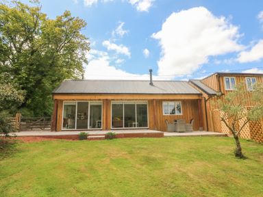A wooden house with sliding glass doors a patio table and chairs and a lawn at Langdale Barn in Sampford Courtenay