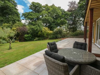 An outdoor patio with a round glass table and four wicker chairs overlooking a grassy yard with trees at Langdale Barn in Sampford Courtenay