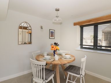A dining area with a round wooden table set with white dishes and four white chairs with cushions near a window with blinds at Kingfisher 2 The Old Stables in Witney