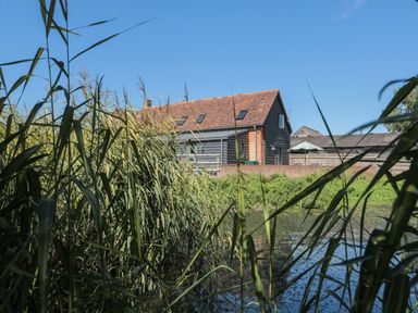 A house with a tiled roof near a pond with tall grasses in the foreground at Kingfisher 2 The Old Stables Witney