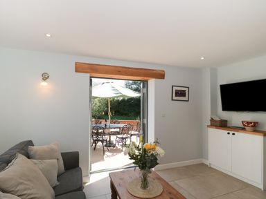 A living room with a gray sofa and cushions a wooden coffee table with flowers and a built in cabinet below a wall mounted television and an open door leading to a patio with a table chairs and an umbrella at Kingfisher 2 The Old Stables in Witney