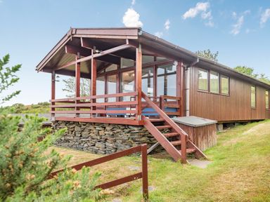 A wooden cabin with a stone foundation and a covered porch with railing and stairs surrounded by grass and bushes at The Look Out in Aberdovey