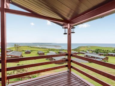 A wooden balcony overlooking cabins green fields and a distant coastline at The Look Out in Aberdovey