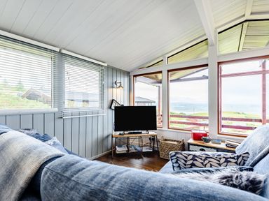A living room with a corner sofa a television on a stand and large windows overlooking a landscape at The Look Out in Aberdovey