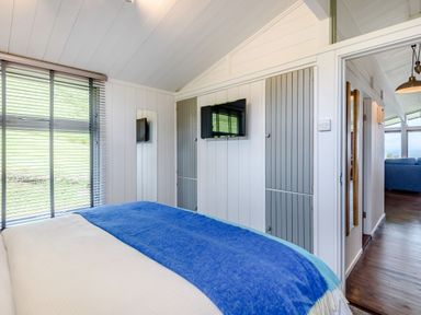 A bedroom with a bed covered in white and blue blankets a wall-mounted TV and large windows with blinds at The Look Out in Aberdovey