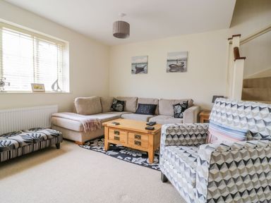 A living room with a beige sectional sofa, a wooden coffee table, a patterned armchair, and paintings of boats on the wall at 26 Burtons Mill in Stalham