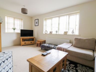 A living room with a beige sofa a wooden coffee table a television on a wooden stand and windows with blinds at 26 Burtons Mill in Stalham