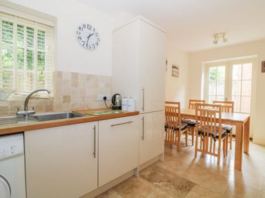 A kitchen and dining area with white cabinets a wooden countertop a sink a clock on the wall a wooden dining table with six chairs and glass doors at 26 Burtons Mill in Stalham