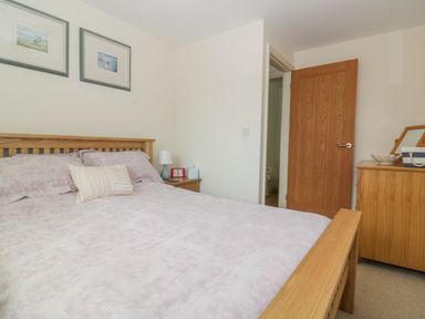 A bedroom with a wooden bed frame a side table with a lamp and clock two framed pictures on the wall a wooden door and a wooden dresser with a mirror at 26 Burtons Mill in Stalham