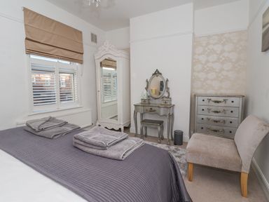 A bedroom with a bed covered in a gray quilt and towels on it a white wardrobe with a mirrored door a vanity table with a mirror and stool a chest of drawers and a cushioned chair at 202 Salisbury Terrace in York