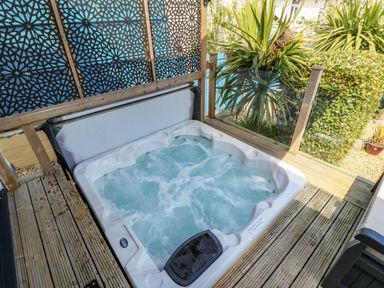 An outdoor hot tub with bubbling water on a wooden deck next to decorative privacy screens and green plants at Daisy Cottage in Benllech