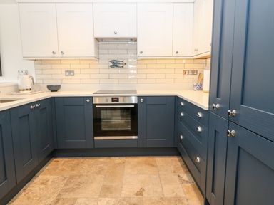 A kitchen with blue lower cabinets and white upper cabinets a built in oven white countertop and tiled backsplash at Arwel in Nefyn