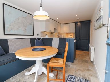 A dining area with a round wooden table and built-in bench seating beside a kitchen with white and dark blue cabinets at Arwel in Nefyn