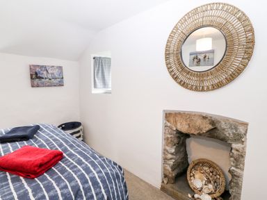 A bedroom with a bed covered with striped blue and white bedding with folded towels a stone fireplace with a basket of shells a round wicker framed mirror and wall art at Arwel in Nefyn