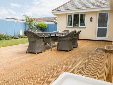 An outdoor wooden deck with a glass table and wicker chairs next to a house with blue fencing at Trevose House in Crantock