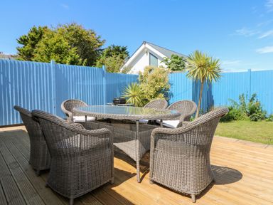 An outdoor patio with a glass top dining table and six wicker chairs on a wooden deck with blue fencing and plants at Trevose House in Crantock