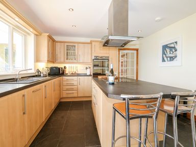 A kitchen with wooden cabinets a black countertop a sink microwave oven and bar stools at Trevose House in Crantock