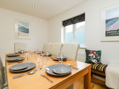 A dining room with a wooden table set with plates glasses and cutlery surrounded by cream chairs and a bench with a cushion and a blanket at Trevose House in Crantock