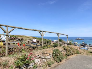 An outdoor seating area with wooden picnic tables near the ocean with parked cars and a small rocky island in the distance at Trevose House in Crantock