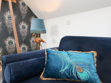 A blue velvet sofa with a decorative peacock feather pillow and a lamp with a flamingo base in a room with peacock feather wallpaper at Trevose House in Crantock