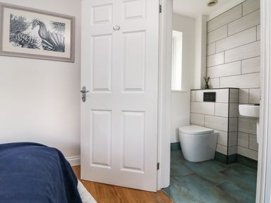 A bedroom with a blue blanket a framed seahorse picture on the wall and an open door leading to a bathroom with a toilet and a small sink at Trevose House in Crantock