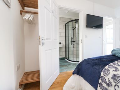A bedroom with a bed a wall-mounted television wooden floor an open door revealing a bathroom with a shower and a closet with hangers at Trevose House in Crantock