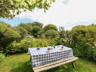 A garden picnic table with a blue and white checkered tablecloth set with tea cups and plates surrounded by trimmed bushes at Higher Roskorwell Cottage in St Keverne