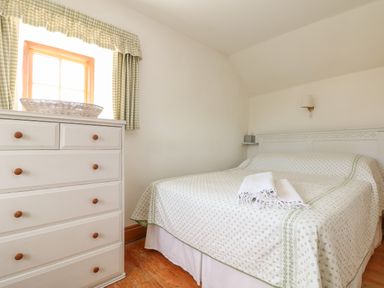 A bedroom with a bed covered in a patterned bedspread next to a white chest of drawers under a window at Higher Roskorwell Cottage in St Keverne