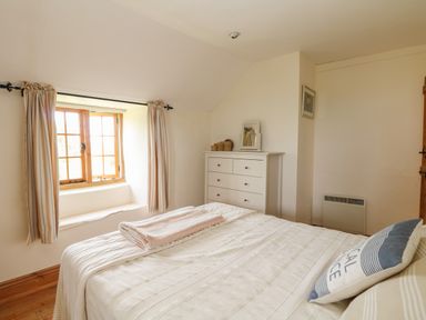 A bedroom with a bed covered with blankets a cushion and a folded towel beside a window with curtains and a chest of drawers at Higher Roskorwell Cottage in St Keverne